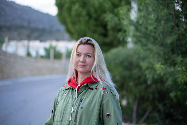 Closeup portrait of a beautiful forty-year-old blonde caucasian woman in a casual green jacket posing outdoors in a park against the background of green trees. - Photo, Image