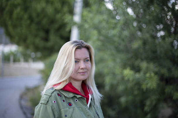 Nahaufnahme Porträt einer schönen vierzigjährigen blonden kaukasischen Frau in lässiger grüner Jacke, die im Freien in einem Park vor dem Hintergrund grüner Bäume posiert. - Foto, Bild
