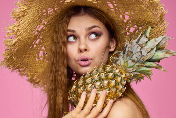 Mujer con un arma en las manos de un sombrero de paja maquillaje brillante frutas exóticas fondo rosa de verano - Foto, Imagen