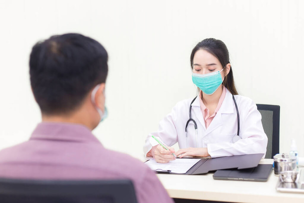 Een Aziatische vrouwelijke arts vraagt naar de ziekte van een mannelijke patiënt. In de onderzoekskamer van het ziekenhuis draagt ze een medisch gezichtsmasker om de ziekte van Coronavirus in het ziekenhuis te voorkomen. Gezondheidszorg, vervuiling PM2.5, nieuw normaal concept. - Foto, afbeelding