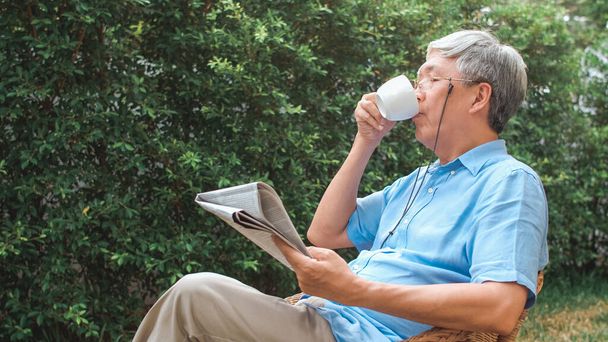 アジア系の先輩たちは自宅でリラックス。アジアのシニア中国人男性は朝のコンセプトで自宅の庭に横たわっている間、休憩時間を楽しむ眼鏡は新聞を読み、コーヒーを飲む. - 写真・画像