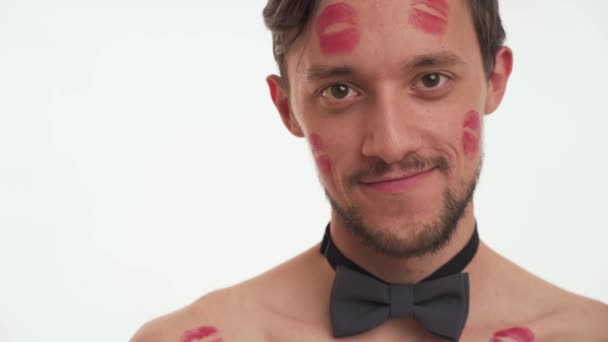 Close up portrait of beautiful young bearded man with brown hair wear grey bow tie with red lipstick marks on face and cheek. One male blowing kiss on white wall background. International Womens day - Footage, Video
