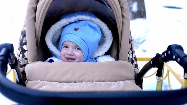 bébé dans un chapeau bleu assis dans une poussette sur une promenade en hiver sourit à maman. enfant gai avec un sourire. - Séquence, vidéo