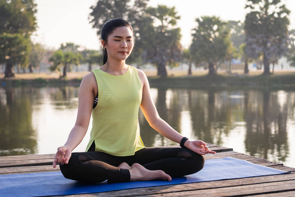 Joven asiática hermosa mujer practicando yoga y meditar en la pose de loto al aire libre junto al lago en la mañana para la relajación y la paz de la mente. Concepto de armonía y meditación. Estilo de vida saludable - Foto, Imagen