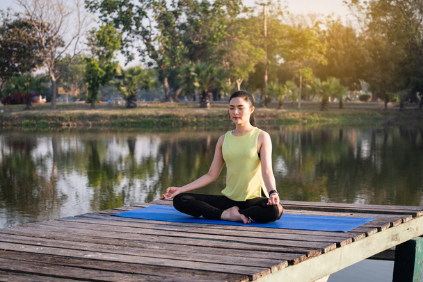 若いアジアの美しい女性ヨガを練習し、蓮のポーズで瞑想心のリラクゼーションと平和のために午前中に湖の横に屋外。調和と瞑想の概念。健康的なライフスタイル - 写真・画像