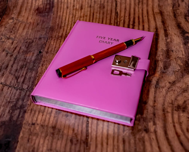 Een roze vijf jaar afsluitbaar dagboek en houten vulpen op een houten achtergrond - Foto, afbeelding