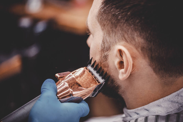 Close-up van kapper met medische handschoenen scheren baard aan man in kapperszaak. Kapsel coronavirus pandemische bescherming - Foto, afbeelding