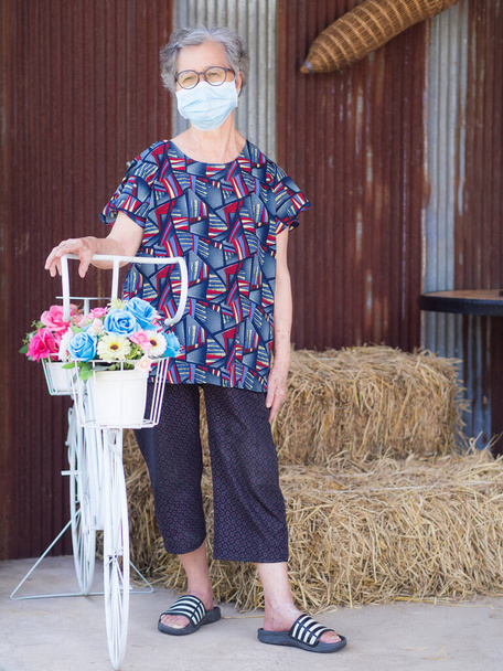 Starší žena s obličejovou maskou, která se dívá do kamery, zatímco stojí u květinového kola. Maska na ochranu viru, koronaviru, pylových zrn. Koncepce věku lidí a zdravotní péče. - Fotografie, Obrázek