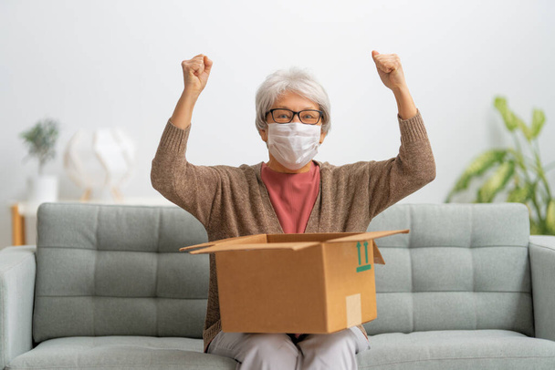 Senior woman is holding cardboard box sitting on sofa at home.  - Foto, Imagem