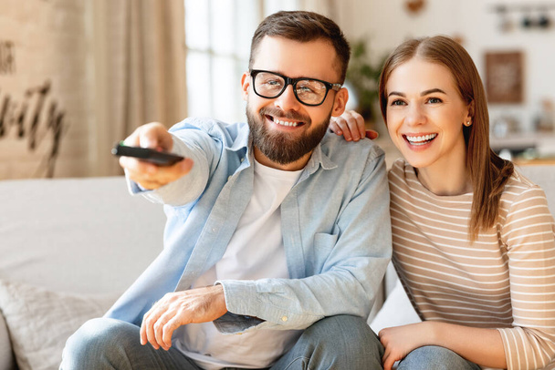 Delighted adult couple: man and woman smiling and looking at camera while sitting on sofa and changing channels on TV with remote control - Fotoğraf, Görsel