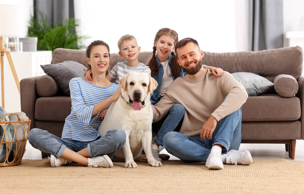 Hymyilevä äiti ja isä pienten lasten kanssa ja ihana puhdasrotuinen labradorinnoutaja koira istuu lattialla ja katselee kameraa viettäessään aikaa yhdessä kotona. - Valokuva, kuva
