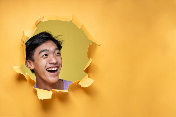 Boldog izgatott fiatal ázsiai tinédzser férfi pózol keresztül szakadt sárga papírlyuk, nézi a másoló hely vagy üres sárga papír, visel lila póló, reklám legjobb termék. Reklámtevékenység. - Fotó, kép