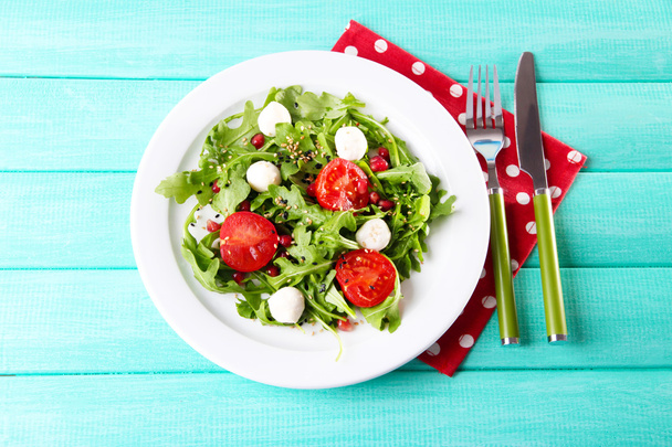 Green salad made with  arugula, tomatoes, cheese mozzarella balls and sesame  on plate, on color wooden background - Photo, Image