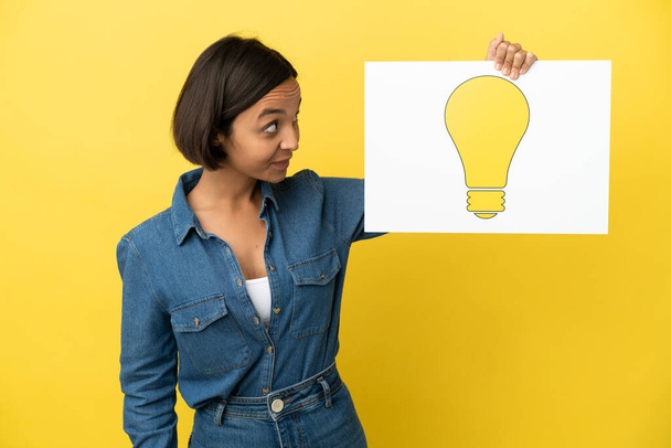 Mladá smíšená rasa žena izolované na žlutém pozadí drží plakát s ikonou žárovky - Fotografie, Obrázek