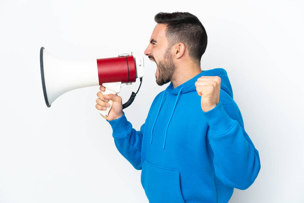 Young caucasian handsome man isolated on white background shouting through a megaphone to announce something in lateral position - Фото, изображение
