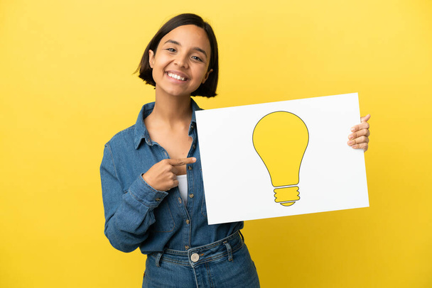 Young mixed race woman isolated on yellow background holding a placard with bulb icon and pointing it - Fotografie, Obrázek