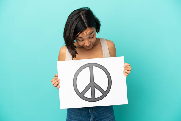 Młoda mieszana rasa kobieta odizolowana na niebieskim tle z tabliczką z symbolem pokoju - Zdjęcie, obraz