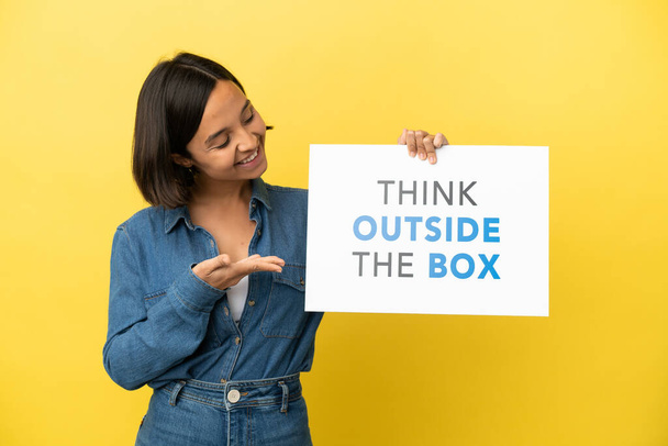 Mujer joven de raza mixta aislada sobre fondo amarillo sosteniendo un cartel con texto Think Outside The Box y señalándolo - Foto, imagen