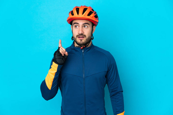 Νεαρός ποδηλάτης απομονωμένος σε μπλε φόντο σκέφτεται μια ιδέα που δείχνει το δάχτυλο προς τα πάνω - Φωτογραφία, εικόνα