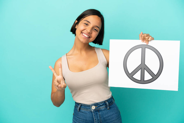 Fiatal vegyes fajú nő elszigetelt kék háttér kezében egy plakát béke szimbólum és ünnepli a győzelem - Fotó, kép