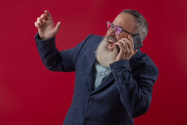 Homem carismático sênior com uma barba cinza em uma camisa leve em uma jaqueta casual escura segurando um smartphone e grita - Foto, Imagem