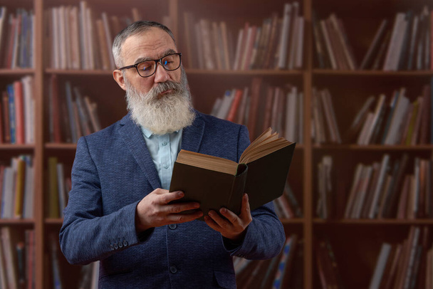 Starszy charyzmatyczny mężczyzna w szarej brodzie w jasnej koszuli w ciemnej kurtce, trzymający książkę w starej bibliotece. - Zdjęcie, obraz