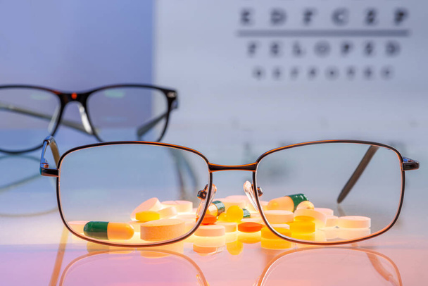 Óculos com lentes e pílulas de prescrição, vitaminas, suplementos dietéticos na mesa de vidro contra um fundo de teste visual com letras em borrão. Conceito de Oftalmologia em Saúde. Foco selecionado - Foto, Imagem