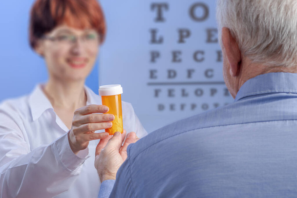 La oftalmóloga saliente le da un frasco de pastillas con receta naranja al hombre mayor de la clínica. Vista desde atrás contra una prueba visual con letras. Enfoque seleccionado en las manos - Foto, imagen