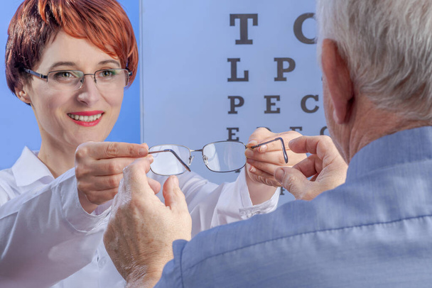 Outgoing female ophthalmologist gives eyeglasses to senior man. View from the back against a visual test with letters. Concept of appointment at the clinic. Selected focus on hands - Photo, Image