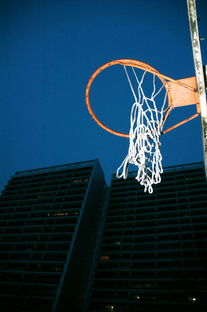 Zgniłe obręcze do koszykówki z rozerwaną siatką na boisku przed symetrycznymi budynkami mieszkalnymi w nocy - Zdjęcie, obraz