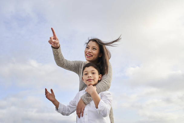 Jeune mère latina pointant vers un certain point tandis que son fils regarde la caméra, contre un ciel bleu vif avec des nuages. Concept de famille. - Photo, image
