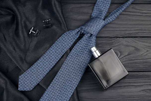 Una botella de colonia para hombre y gemelos con corbata azul se encuentran sobre un fondo de tela de lujo negro sobre una mesa de madera. Accesorios clásicos para hombre. DOF poco profundo - Foto, imagen