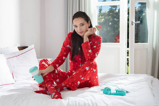 The young, beautiful, brunette girl, in bed and in her red pajamas, poses with a colorful drink and candy in her hand, smiling for the camera. It's a sunny day and the attractive teen age girl - Photo, Image