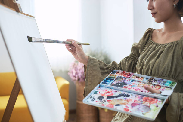 Eine asiatische Frau, die Kunst liebt oder eine weibliche Künstlerin hält eine Farbpalette und Pinsel in der Hand, während sie zu Hause fröhlich auf weißem Papier malt. - Foto, Bild