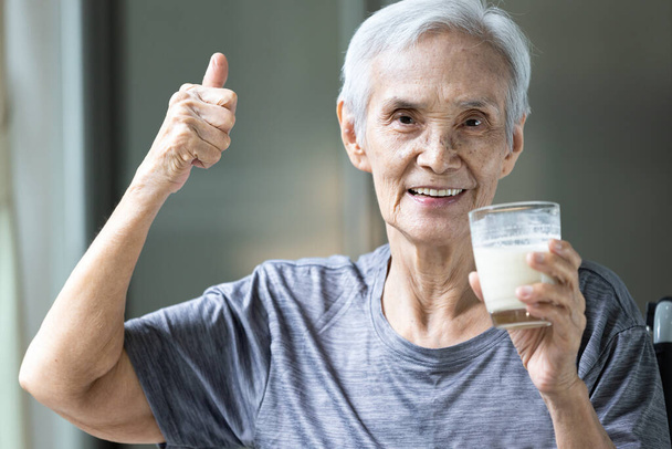 Gesunde alte Großmutter streckt beim Frühstück den Daumen nach oben für leckere frische Milch, glücklich lächelnde asiatische Seniorin trinkt warme Milch oder Nahrungsergänzungsmittel aus dem Glas, Gesundheitsfürsorge - Foto, Bild