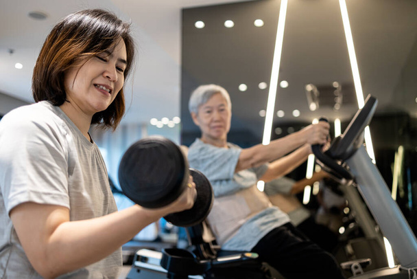 Asian woman working out with dumbbell,physical exercise by lifting weights,old elderly sitting on the spinning bicycle,exercise together,daily workouts in the gym,healthy lifestyle,health care concept - Photo, Image