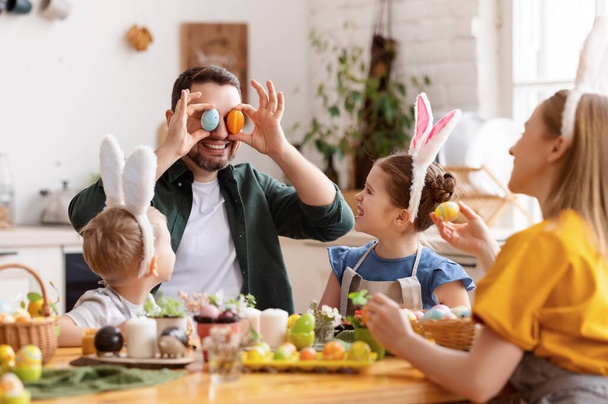 Padre juguetón cubriendo los ojos con huevos de Pascua y sonriendo mientras se reúnen con la familia en la mesa en la cocina moderna juntos - Foto, imagen