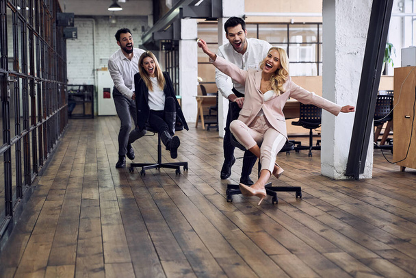 Трудитесь усердно. Четыре молодых веселых бизнесмена в формальной одежде веселятся, гоняясь на офисных стульях и улыбаясь - Фото, изображение