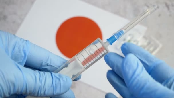 injectiespuit met een covid-19 vaccin wordt met de hand vastgehouden in een handschoen op de achtergrond van de Japanse vlag, vaccin tegen coronavirus, vaccinatie in Japan start concept - Video