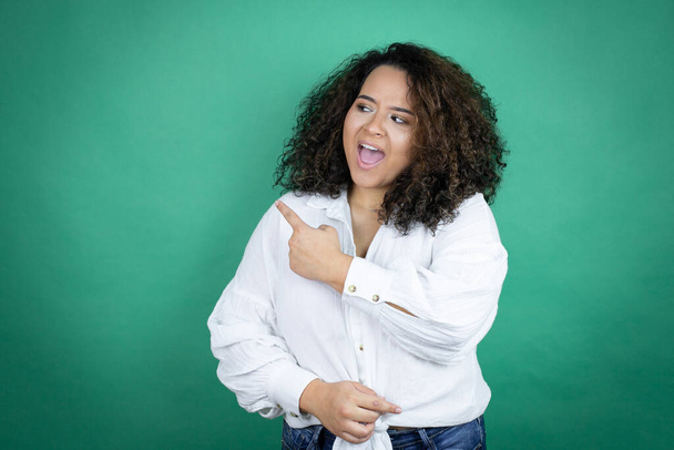 Giovane ragazza afroamericana indossa camicia bianca su sfondo verde sorridente e puntando con mano e dito di lato - Foto, immagini