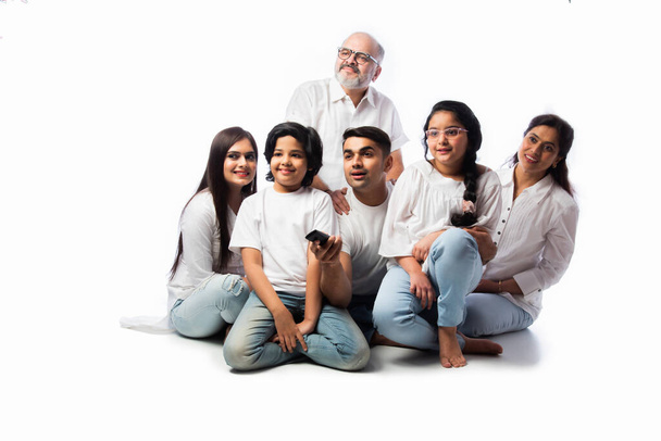 Indian asian multigenerational family of six watching TV together at home and having fun , sitting on floor against white background - Photo, Image