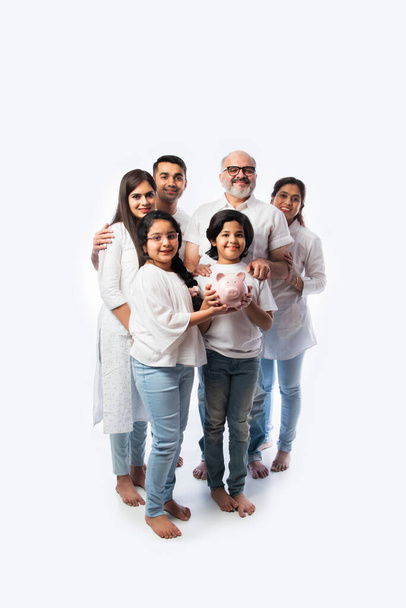 Indische Mehrgenerationenfamilie mit sechs Kindern hält Sparschwein, während sie weiße Tücher trägt und vor weißer Wand steht - Foto, Bild