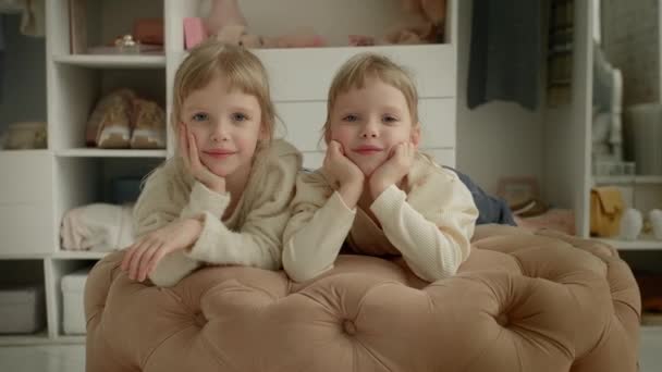 Vidéo 4K de deux élégantes petites sœurs jumelles filles souriantes sur tabouret rembourré ensemble dans le dressing sur fond de garde-robe - Séquence, vidéo