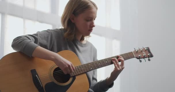 Підліток грає на гітарі в білій вітальні з красивим сонячним світлом. Жінка створює музику в приміщенні. Дівчина практикує музичний інструмент сама під час пандемії
. - Кадри, відео