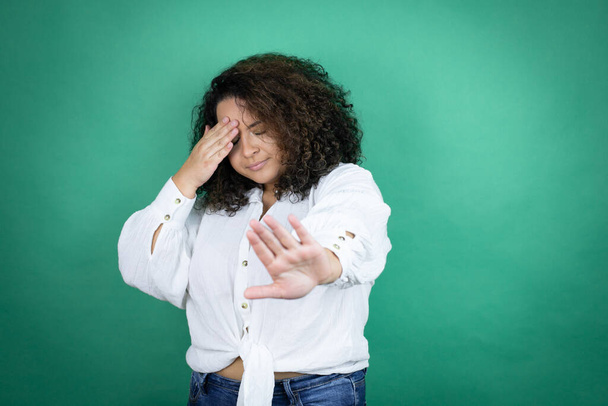 Молода афроамериканська дівчина, одягнена в білу сорочку на зеленому фоні, закриває очі руками і припиняє жести з сумом і страхом вираження. - Фото, зображення
