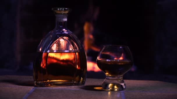 Bottiglia e bicchiere con whisky o cognac sullo sfondo del fuoco nel camino - Filmati, video