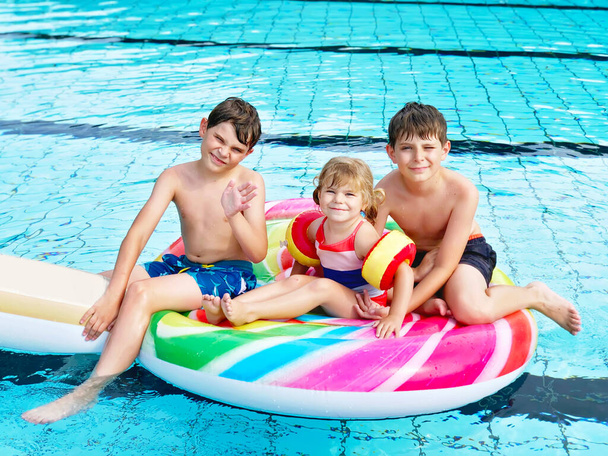 Tres niños, dos niños y una niña chapotean en una piscina al aire libre en verano. Niños felices, hermanos y hermanas jugando en una enorme piruleta inflable de chicle disfrutando de un clima soleado en la piscina pública. - Foto, Imagen