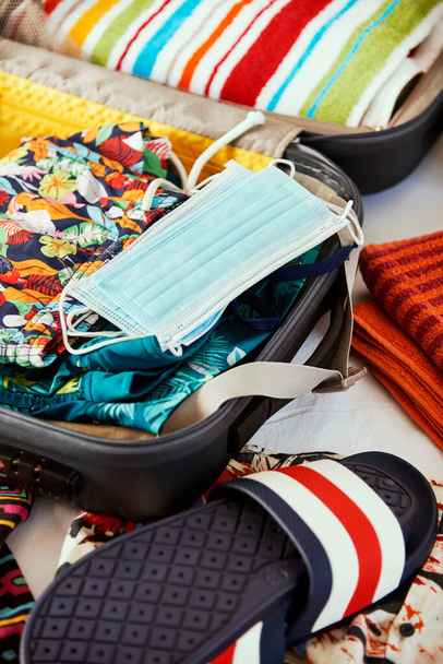 close-up de algumas novas máscaras descartáveis em uma mala embalada com alguns itens de férias diferentes, como toalhas de praia, calções de banho ou sandálias de praia - Foto, Imagem