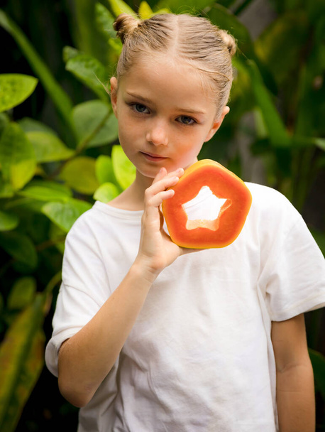 Taze papayası olan sevimli küçük bir kız. Kafkasyalı bir kız boynunun önünde papaya meyvesi tutuyor. Seçili odak. Yeşil tropik yapraklar arka planda. Organik meyve konsepti. Bali, Endonezya - Fotoğraf, Görsel