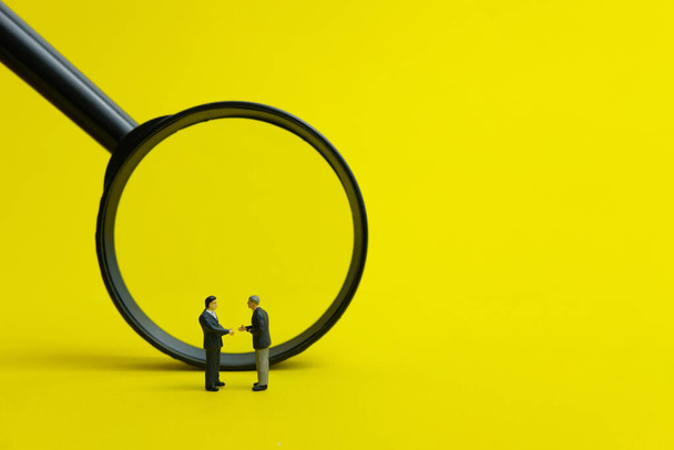 Miniatuur mensen, partnerschap concept - twee zakenman maken een handdruk voor vergrootglas met gele achtergrond - Foto, afbeelding